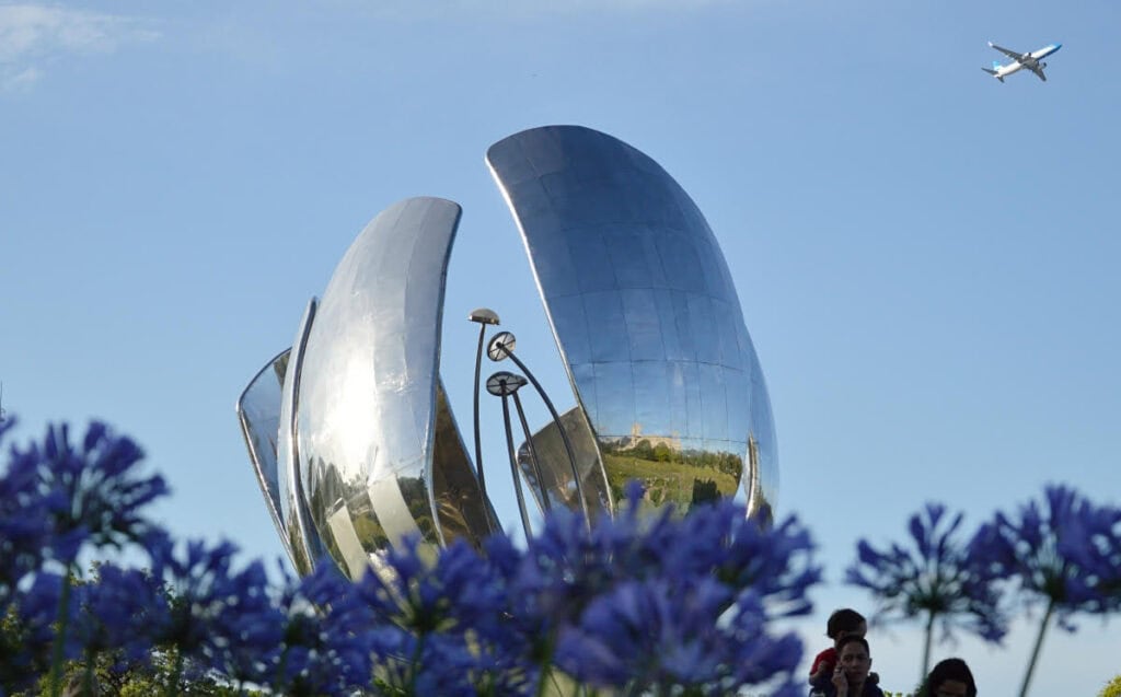 This image features the Floralis Genérica, a famous steel sculpture located in Buenos Aires, Argentina. The large metallic flower reflects the sky and nearby surroundings, with purple flowers in the foreground and a clear blue sky above. An airplane is visible in the top right corner of the image, adding a sense of motion to the otherwise calm scene. People can be seen near the bottom right, enjoying the view of this landmark.