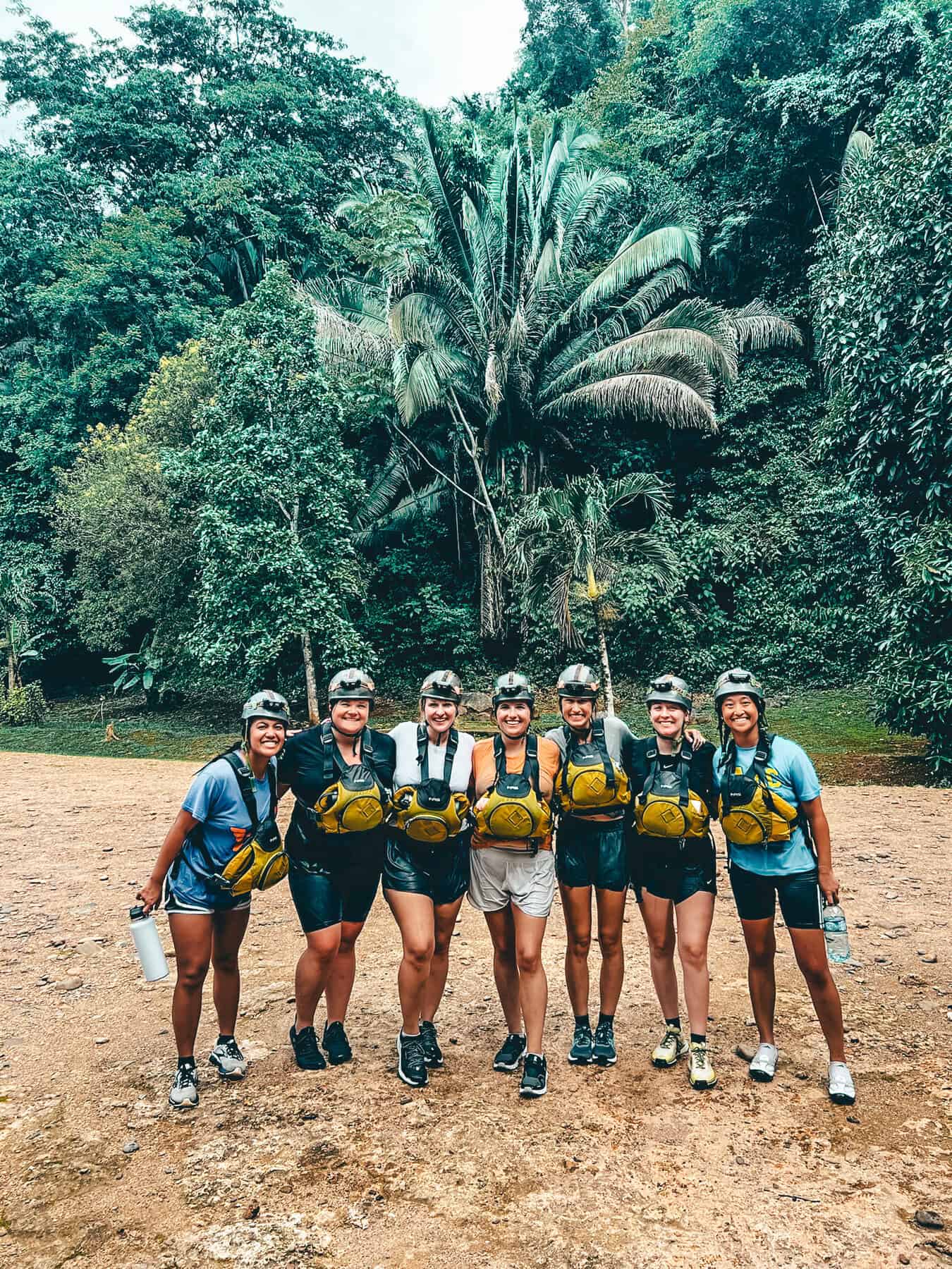 A group of seven people poses together in front of lush jungle foliage, wearing helmets, life jackets, and athletic attire, suggesting they are about to embark on an outdoor adventure. The setting is a natural area with a mix of palm trees and dense greenery, likely preparing for a tour of the ATM Cave in Belize.