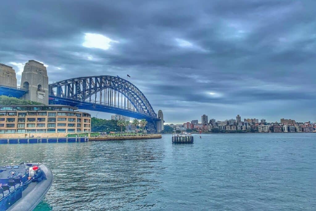 This image features the iconic Sydney Harbour Bridge in Australia, spanning over the harbor with the city's skyline in the background. The overcast sky adds a dramatic feel to the scene, while the calm water reflects the surrounding buildings and structure. A nearby building and a small boat in the foreground create a sense of activity around the harbor, and the bridge's impressive steel arches stand out as a key architectural feature. The setting showcases a blend of urban life and natural waterfront beauty.