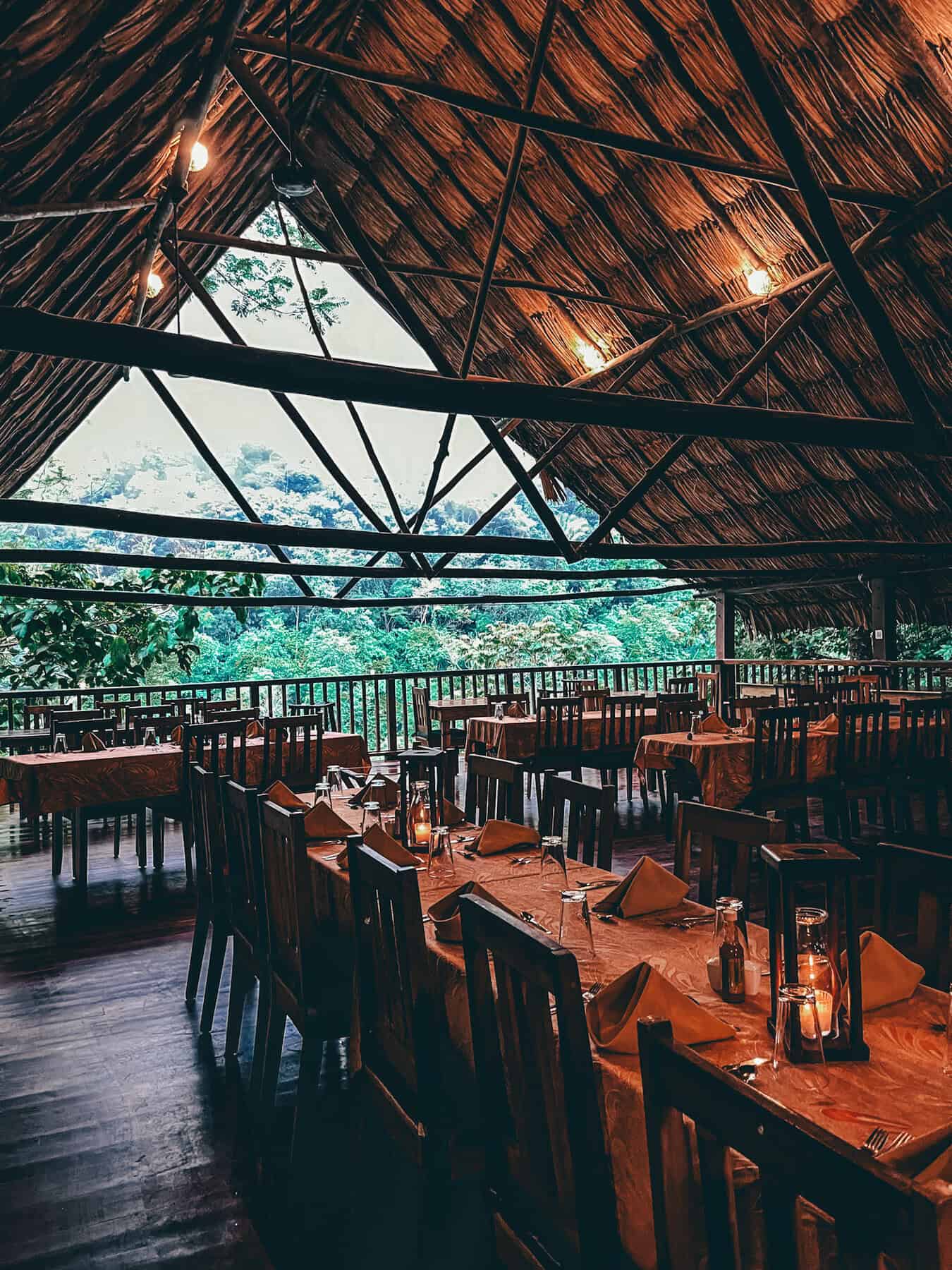 A rustic, open-air restaurant with a thatched roof and wooden beams overlooks lush greenery at Black Rock Lodge. The tables are elegantly set with folded napkins, glassware, and lanterns, creating a cozy ambiance with soft lighting.