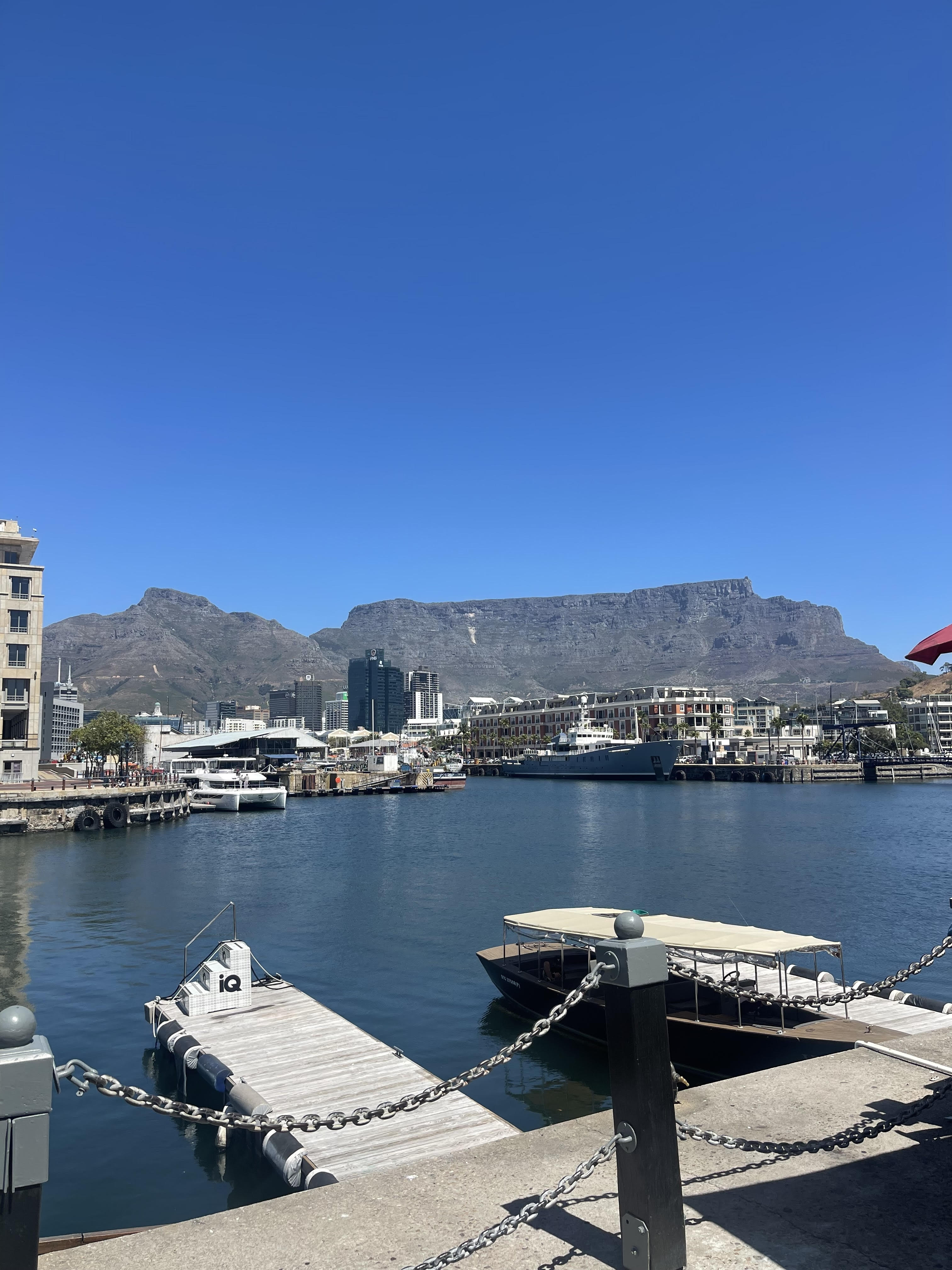 This image shows the scenic Victoria & Alfred Waterfront in Cape Town, South Africa, with Table Mountain prominently visible in the background under a clear blue sky. The calm water of the harbor reflects the surrounding buildings and boats docked at the piers. A small boat is moored in the foreground, and modern buildings line the waterfront, with a backdrop of the iconic flat-topped mountain. The image captures a serene yet vibrant urban setting at the foot of a natural landmark.