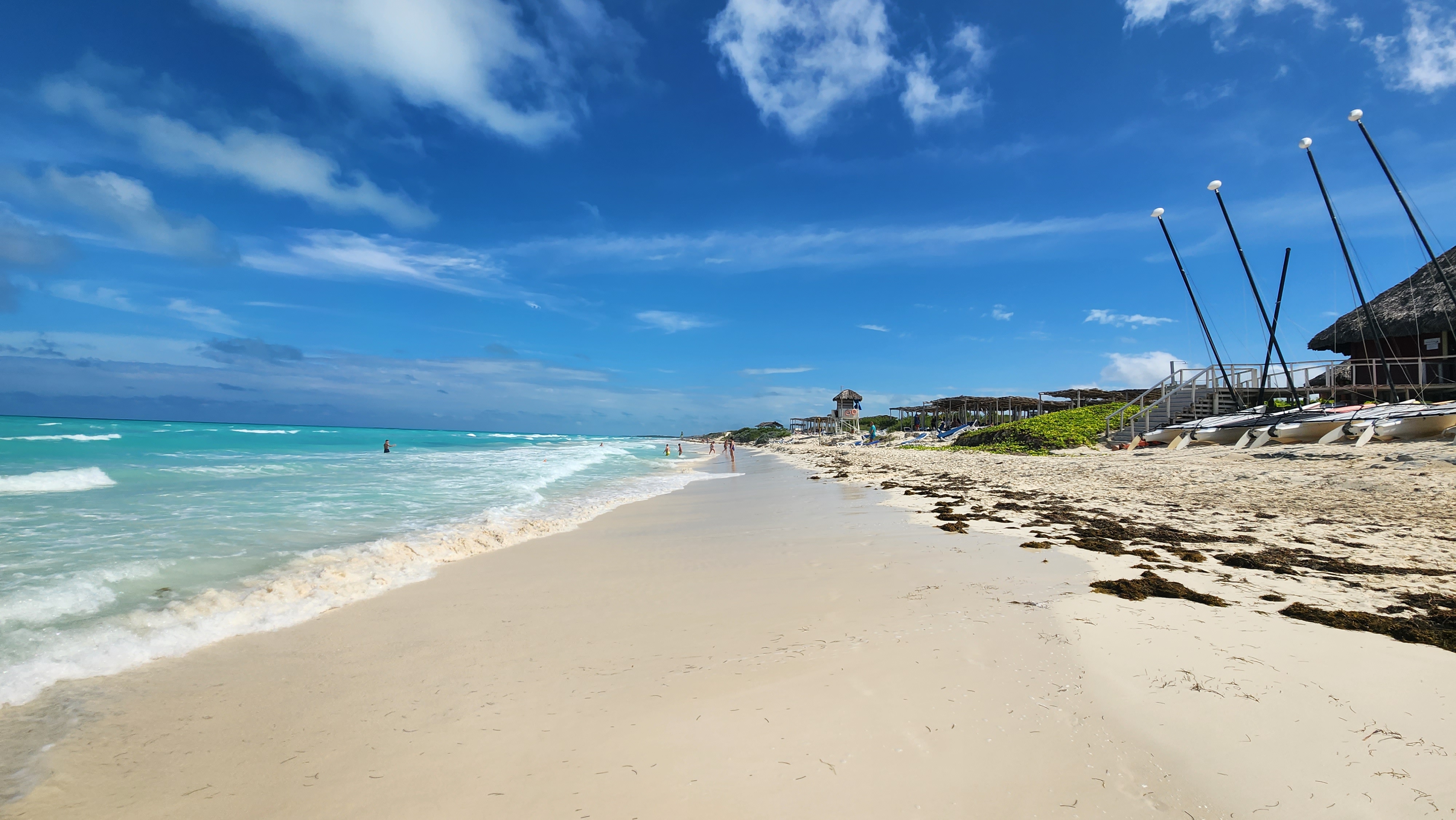 This image shows a tranquil beach in Cuba with soft white sand stretching along the shoreline. The turquoise water gently meets the sand, with a few people wading in the distance under a bright blue sky with scattered clouds. To the right, beach huts and sailboats, lined up with their masts reaching skyward, create a relaxed and inviting atmosphere. The beach appears quiet and pristine, offering a peaceful seaside experience.