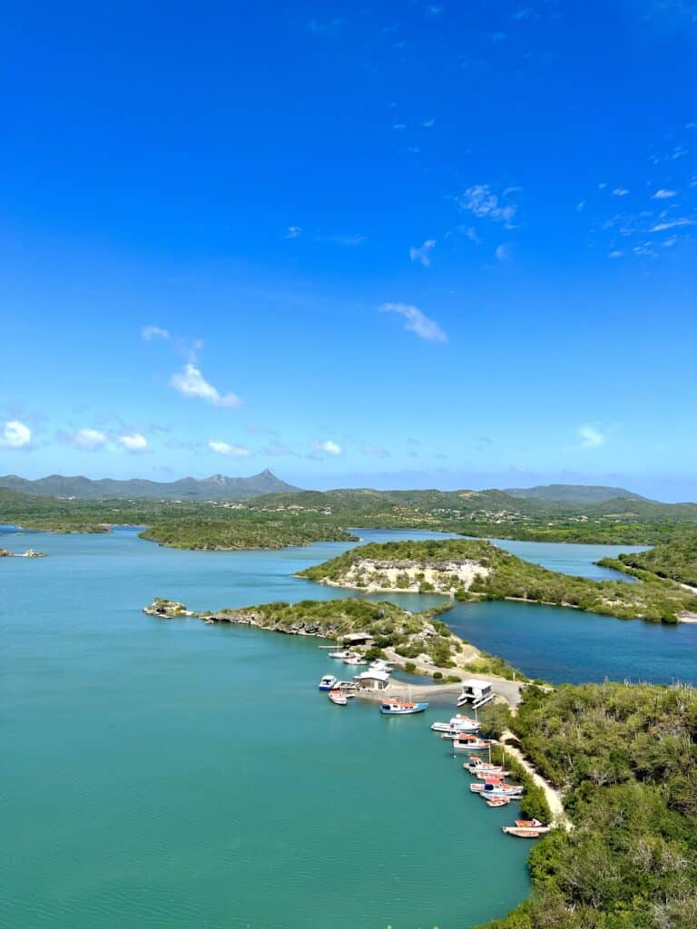 This image showcases a beautiful aerial view of a coastal area in Curaçao, with small islands and boats docked along the shore in the foreground. The clear turquoise water contrasts with the lush green vegetation that covers the land. The distant landscape features rolling hills under a vibrant blue sky with a few scattered clouds. The peaceful setting and scenic coastline capture the tropical charm and natural beauty of Curaçao.