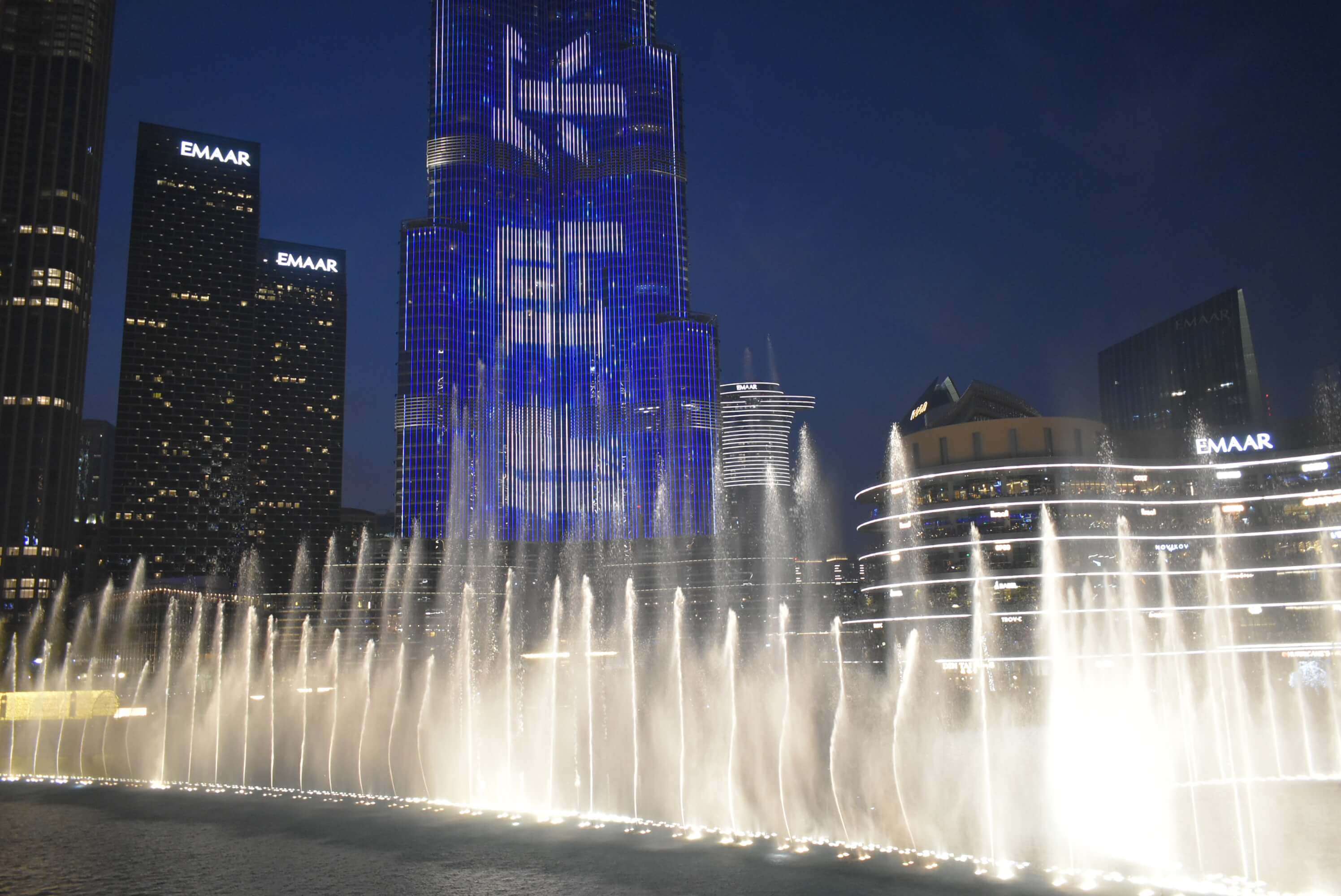 This image captures the Dubai Fountain in action at night, with water jets illuminated and dancing in front of the towering Burj Khalifa, which is lit up with vibrant blue and white light displays. Surrounding skyscrapers, such as the Emaar buildings, are also lit, contributing to the city's iconic nighttime skyline. The bright lights of the fountain and the surrounding buildings reflect on the water, creating a dynamic and awe-inspiring scene in the heart of Dubai, UAE.