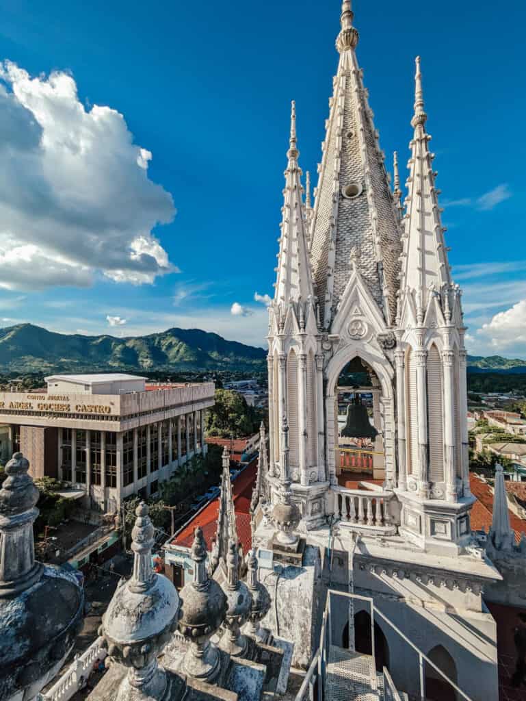 This image captures a stunning view of a neo-Gothic church tower, likely part of the Cathedral of Santa Ana in El Salvador. The intricate spire stands tall against a clear blue sky, with detailed stonework and a bell visible inside the structure. In the background, you can see lush green mountains and the modern architecture of a nearby judicial building, creating a contrast between historic and contemporary elements. The angle of the shot highlights the grandeur of the cathedral’s design and the scenic landscape beyond.