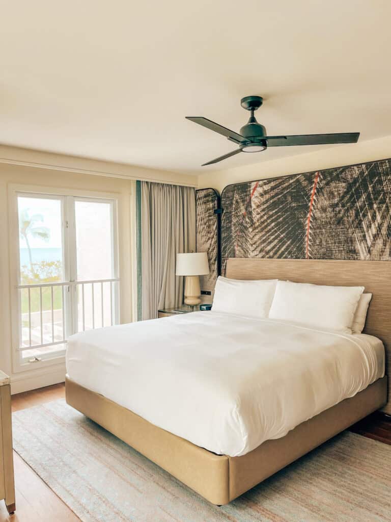 The first image shows a serene bedroom with a large bed, white linens, and a ceiling fan, accented by a textured wall with palm imagery. Soft lighting and a view through a glass door reveal a tropical landscape, enhancing the room’s calm and luxurious vibe.