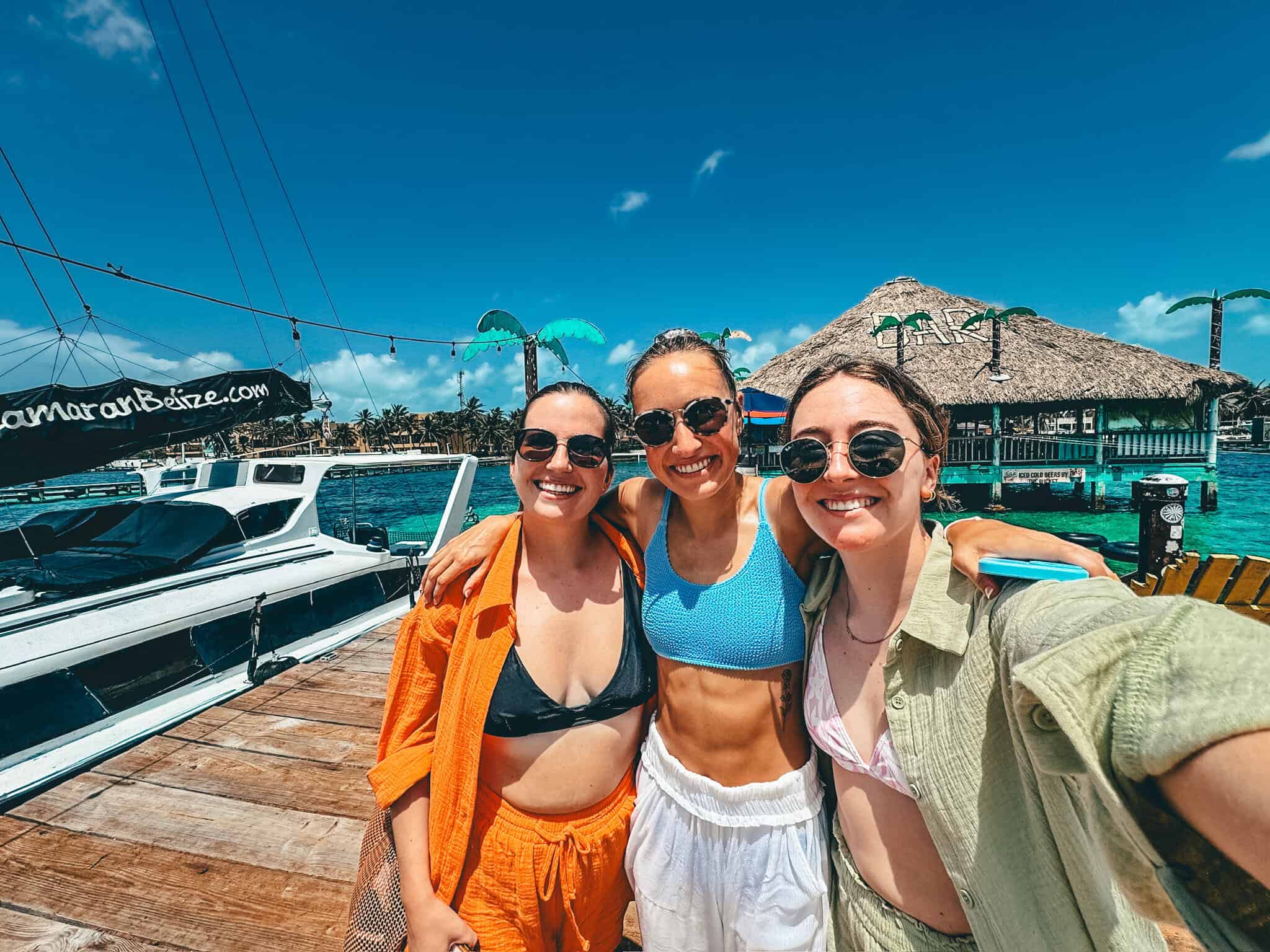 Three friends pose for a selfie on a sunny dock at Caye Caulker, Belize, with a catamaran and a thatched-roof bar in the background. They are dressed in beachwear and sunglasses, smiling against a backdrop of turquoise water and clear blue skies.