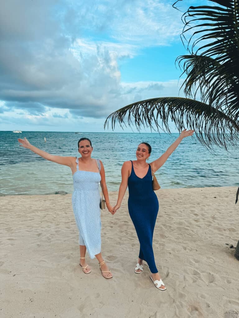 Two friends holding hands and smiling on a sandy beach in San Pedro, Belize. They’re dressed in summery blue dresses, standing under a partly cloudy sky near the ocean