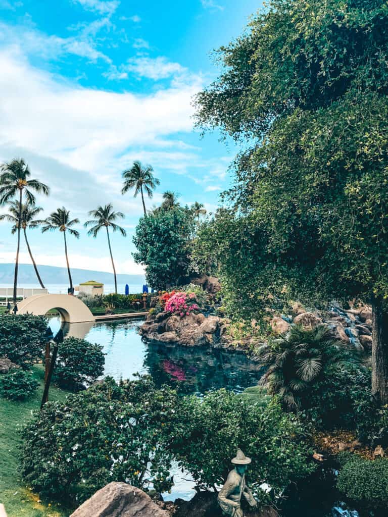 This image showcases a tranquil garden at the Hyatt Regency Maui, featuring a small pond surrounded by lush tropical plants and a variety of greenery. A quaint arched bridge crosses over the water, while a few bright pink flowers and tiki torches add visual accents. In the background, tall palm trees and a glimpse of distant mountains complete the serene, island landscape.