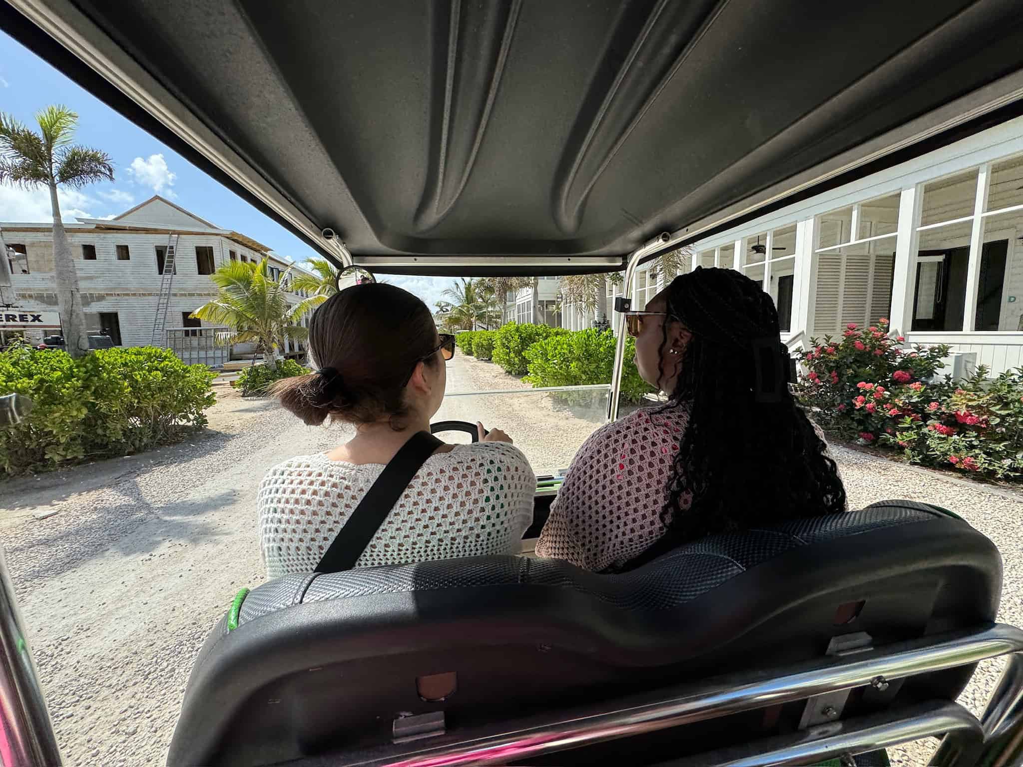 Two women ride in a golf cart along a dirt road lined with tropical plants and white buildings under a sunny sky. The view is from the back seat, capturing the relaxed atmosphere and a mix of greenery and construction in the background.