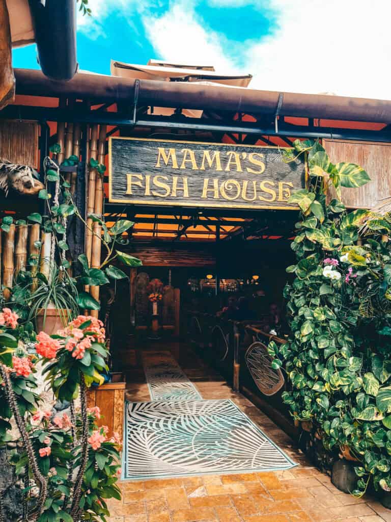 The image shows the entrance to Mama's Fish House, a well-known seafood restaurant in Maui, Hawaii. The entrance is adorned with lush greenery, vibrant tropical flowers, and a rustic wooden sign with gold lettering reading "Mama's Fish House." The scene invites visitors into a cozy, tropical ambiance under a bright blue sky.