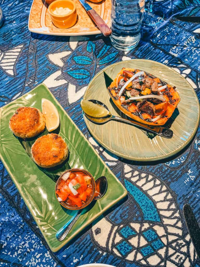 The image showcases a vibrant appetizer spread on a tropical-themed tablecloth. On the left, a green platter holds two golden, crispy cakes with a wedge of lime and a small dish of fresh salsa. On the right, a roasted half papaya is filled with grilled vegetables and topped with herbs, accompanied by a lime wedge. The presentation and colorful ingredients evoke a tropical, island-inspired cuisine.