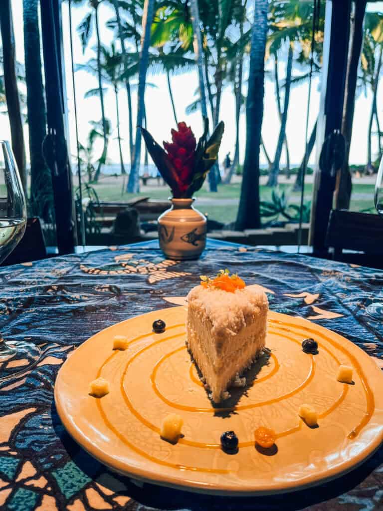 The image displays a slice of tropical dessert, likely coconut cake, artfully plated with a swirl of sauce and garnished with small pieces of fruit like blueberries and pineapple. The dessert is set on a vibrant, patterned tablecloth with a view of a lush palm grove and ocean in the background, complemented by a red tropical flower in a small vase, creating a serene tropical dining ambiance.