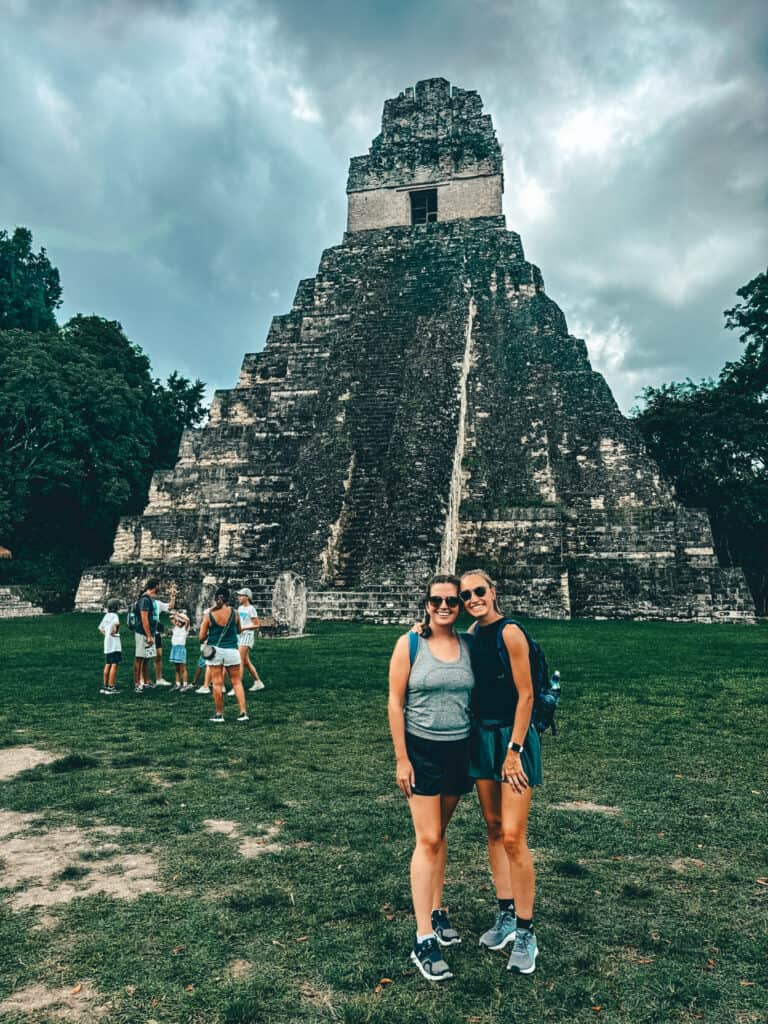 Two friends posing in front of an ancient Mayan pyramid, with a towering stone structure against a dramatic cloudy sky. They are dressed casually in shorts, tank tops, and sneakers, surrounded by lush greenery and other visitors exploring the site.