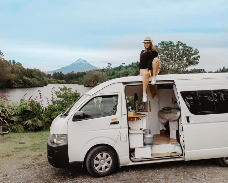 This image shows a woman sitting on top of a white camper van parked in a scenic outdoor location, with lush greenery and a serene river nearby. In the distance, a snow-capped mountain, likely Mount Taranaki in New Zealand, rises against a calm sky, adding to the picturesque landscape. The open van door reveals a cozy interior setup, suggesting a van life adventure in a beautiful natural setting. The woman, dressed casually with a hat, exudes a sense of relaxation and exploration.