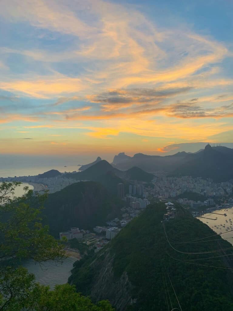 This image shows a stunning sunset view over Rio de Janeiro, Brazil, with the iconic Sugarloaf Mountain and surrounding cityscape in the foreground. The sky is painted with soft hues of yellow, orange, and blue as the sun sets behind the distant mountains. The lush green hills and winding coastline, including famous beaches like Copacabana, can be seen below, along with the city's buildings nestled between the natural landscape. The serene beauty of the scene highlights Rio's blend of urban life and natural splendor.