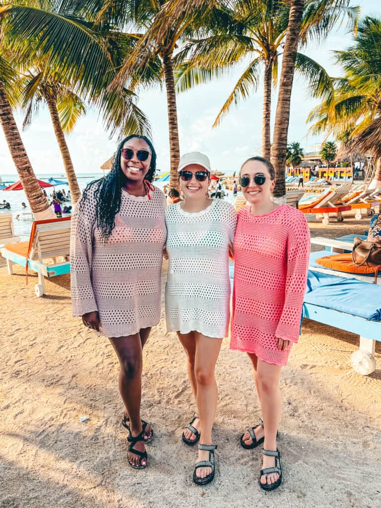 Three friends stand together on a sandy beach, wearing crochet cover-ups and sandals, with palm trees and lounge chairs in the background. The beach scene is lively, with colorful umbrellas and people enjoying the water under a bright sky at what appears to be Secret Beach in Belize.