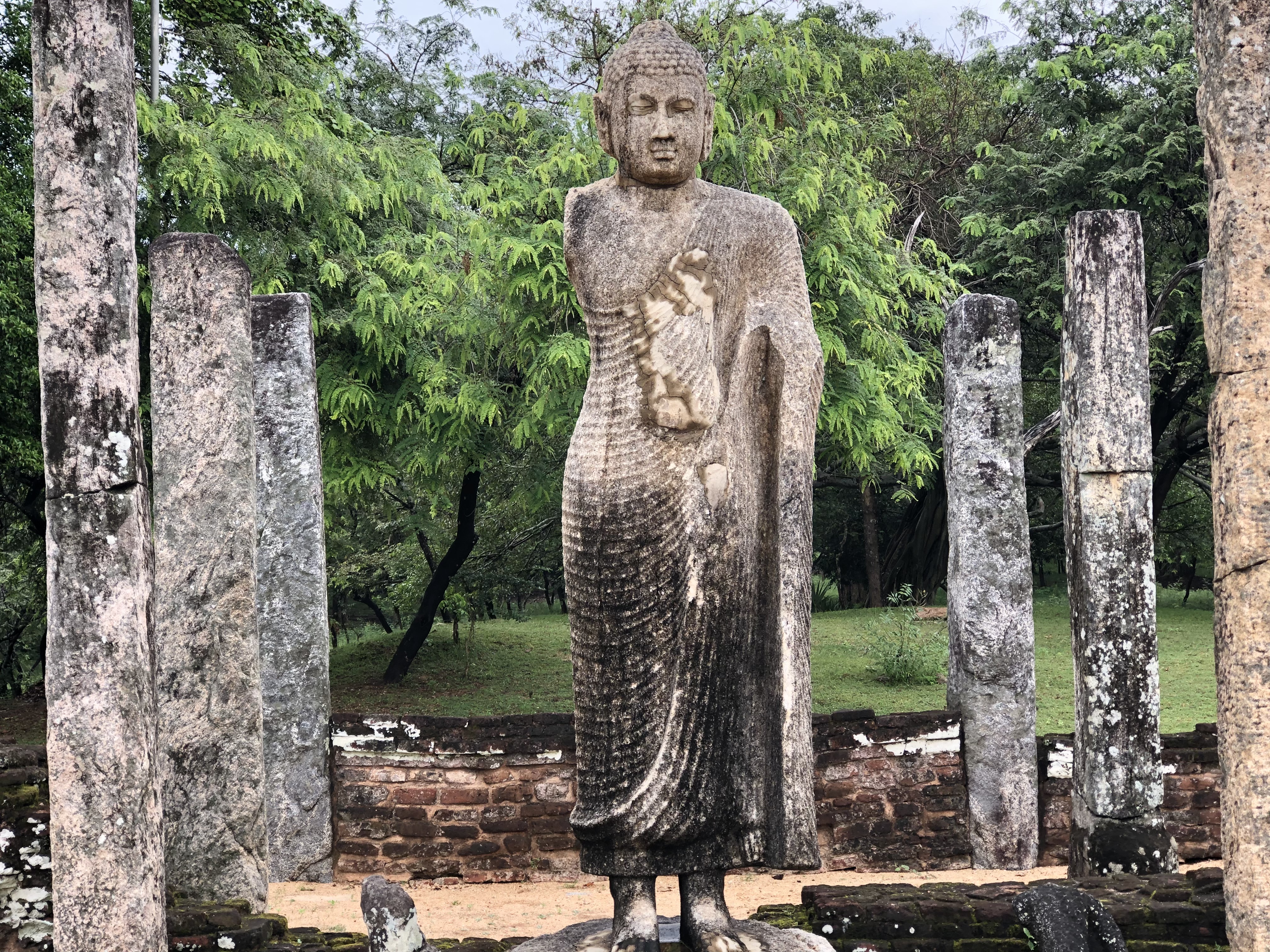 This image shows an ancient stone Buddha statue standing in the ruins of an old temple site in Sri Lanka. The statue is surrounded by weathered stone pillars, with lush green trees in the background, indicating a serene and natural setting. The sculpture, though worn by time, retains its peaceful expression and intricate carvings of robes, symbolizing the rich cultural and religious history of the region. The atmosphere feels tranquil, blending history with the beauty of nature.