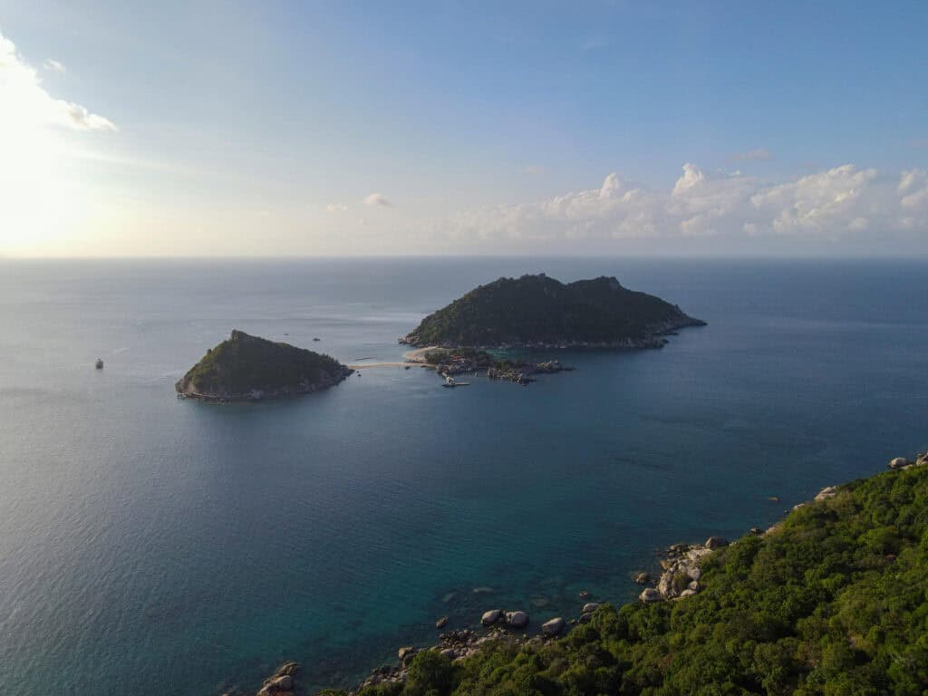 This image showcases a tranquil aerial view of two small islands surrounded by calm, blue waters, likely located off the coast of Thailand. The islands are covered in lush green vegetation, and a small sandy beach connects the two. The serene sea stretches out into the horizon under a soft, pastel sky with light clouds, creating a peaceful and picturesque tropical scene. The sunlight, either from early morning or late afternoon, adds a gentle warmth to the landscape.