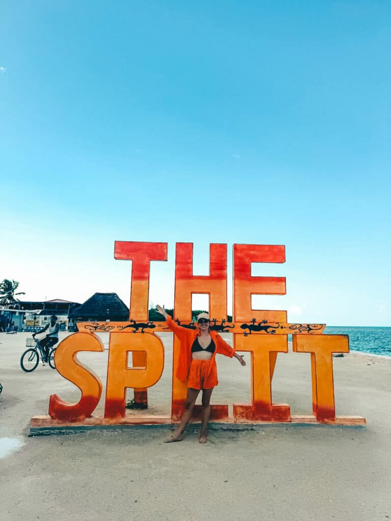 A woman stands barefoot in front of the large, colorful "The Split" sign at Caye Caulker, Belize, wearing a black bikini and an orange cover-up. The beach setting features a bright blue sky, with the ocean visible to the right and a few people and huts in the background.