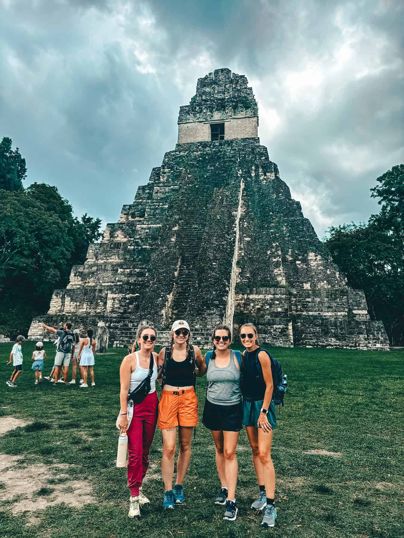 Four women pose in front of the ancient Temple I pyramid at Tikal, Guatemala, on a cloudy day. The structure’s steep steps and stone facade are visible behind them, surrounded by lush greenery, while other tourists explore in the background.