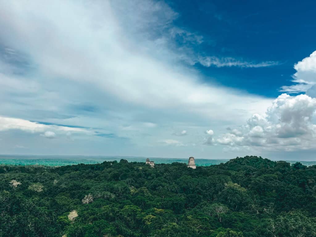 A scenic view of the Tikal Mayan ruins in Guatemala, with ancient temple tops rising above the dense jungle canopy. The landscape stretches into the distance under a sky filled with a mix of clouds and blue, highlighting the expansive and lush surroundings.