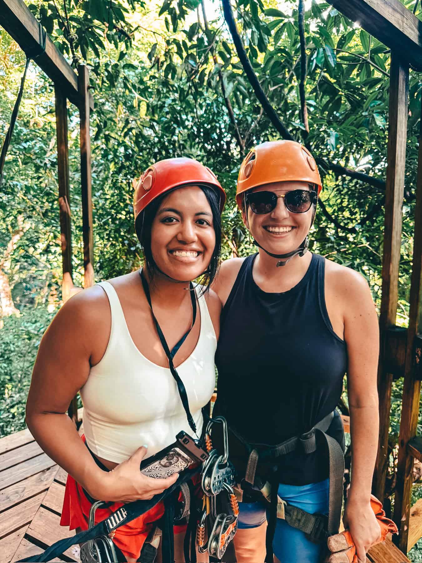 Two women wearing helmets and harnesses smile for the camera during a ziplining adventure. They are standing on a wooden platform surrounded by lush greenery, ready for the outdoor activity.