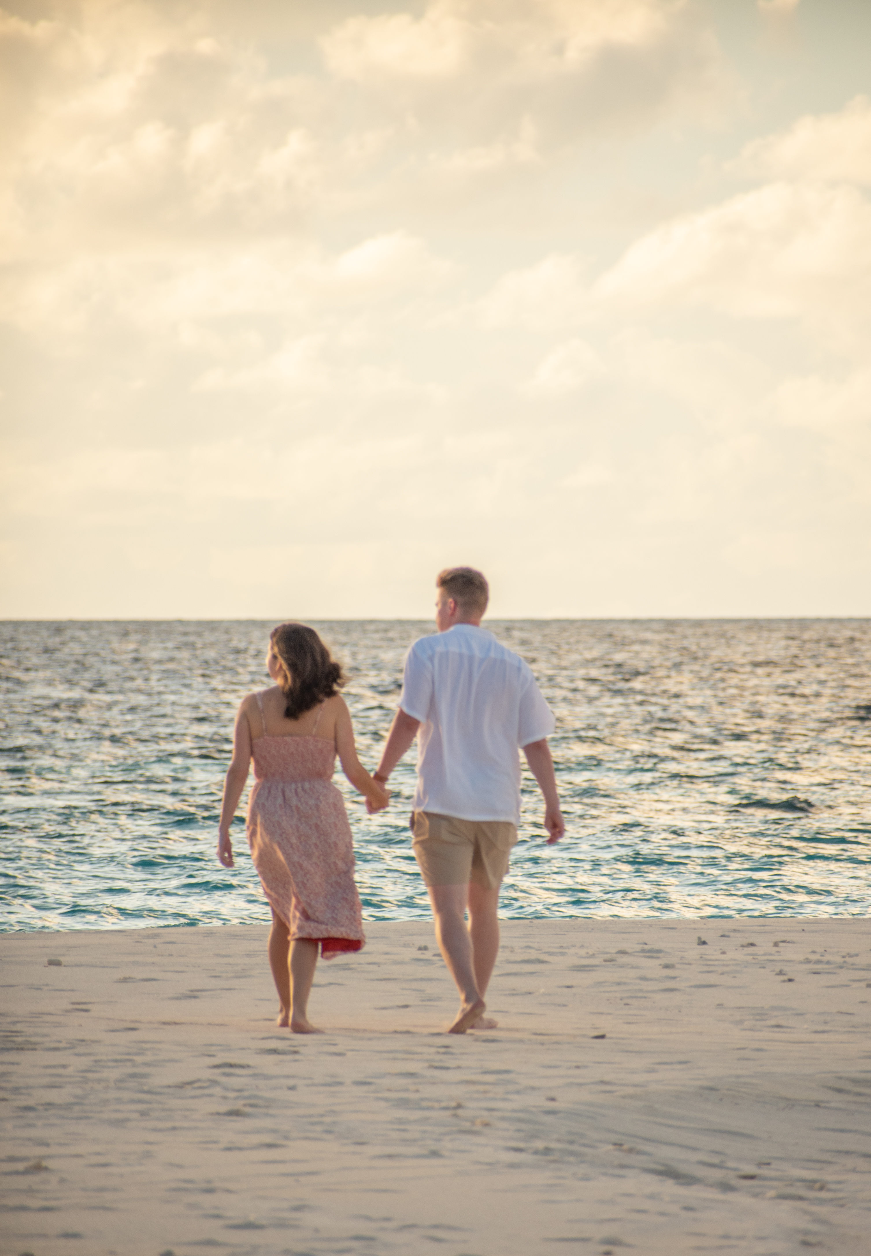 A couple walks hand in hand along a sandy beach, facing the ocean under a sky filled with soft clouds. The warm light suggests a peaceful sunset or early evening, adding a romantic ambiance to their quiet stroll by the water.