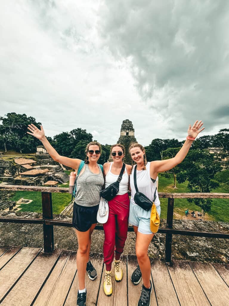 Three friends stand on a wooden platform with lush greenery and ancient stone structures of Tikal, Guatemala, visible in the background. They smile and pose with their arms raised, radiating excitement as they explore the historic Mayan ruins under a cloudy sky. Their casual, adventure-ready attire and backpacks suggest they are prepared for a day of hiking and sightseeing.