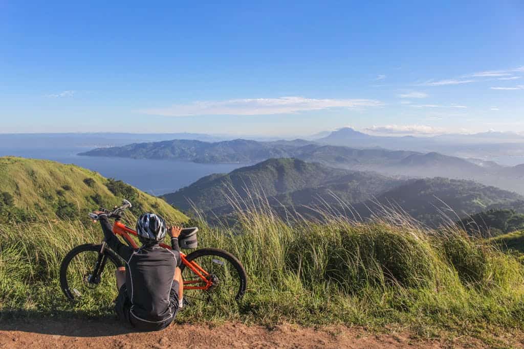 A mountain biker resting by their bike, enjoying a scenic view of lush hills and distant water under a bright blue sky.