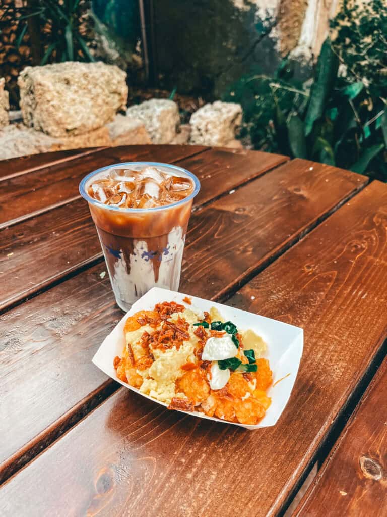 This image features a serving of breakfast totchos—crispy tater tots topped with scrambled eggs, shredded cheese, green onions, crumbled bacon, and a dollop of sour cream—in a white paper tray. Beside it is a clear plastic cup filled with iced coffee or cold brew, swirled with cream. Both items are placed on a wooden table with a rustic stone and greenery backdrop.