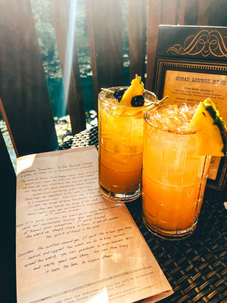 This image shows two tropical cocktails served in clear, ice-filled glasses, each garnished with a pineapple wedge and dark cherries. The drinks are placed on a woven black table, alongside a menu from the "Nomad Lounge" and a note welcoming travelers. Sunlight streams through wooden railings in the background, creating a warm, inviting atmosphere.