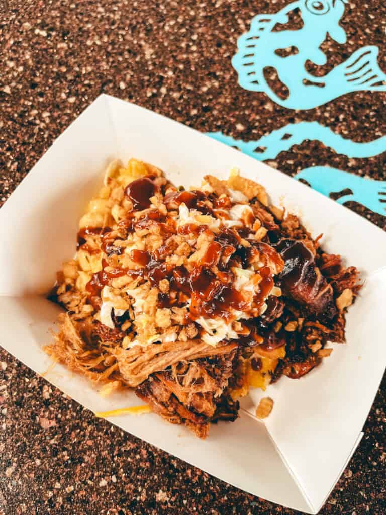 This image shows a hearty serving of pulled pork mac and cheese in a white paper food tray. The dish is topped with crispy fried onions, creamy coleslaw, and a generous drizzle of barbecue sauce. It’s set on a speckled brown table with a blue fish graphic visible in the background.