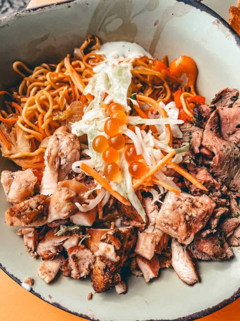 This image shows a colorful noodle bowl featuring a mix of grilled chicken chunks, sliced beef, and stir-fried noodles. The dish is topped with shredded cabbage and carrots, bright orange fish roe, and a drizzle of creamy white sauce, all served in a rustic, light green bowl with a dark rim.