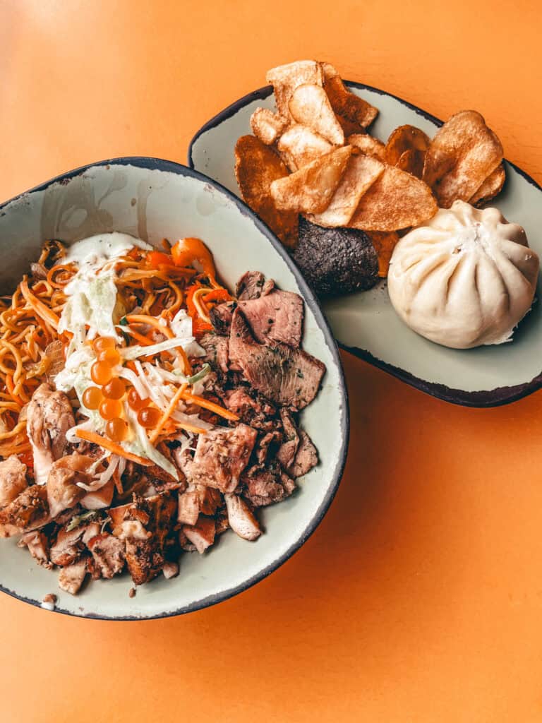 This image shows a meal consisting of a vibrant noodle bowl and a side plate on an orange table. The noodle bowl features grilled chicken, sliced beef, stir-fried noodles, shredded cabbage and carrots, topped with fish roe and creamy sauce. The side plate includes crispy potato chips, a steamed bao bun, and a dark-colored sausage or meat item. Both dishes are served in rustic, light green bowls with dark rims.