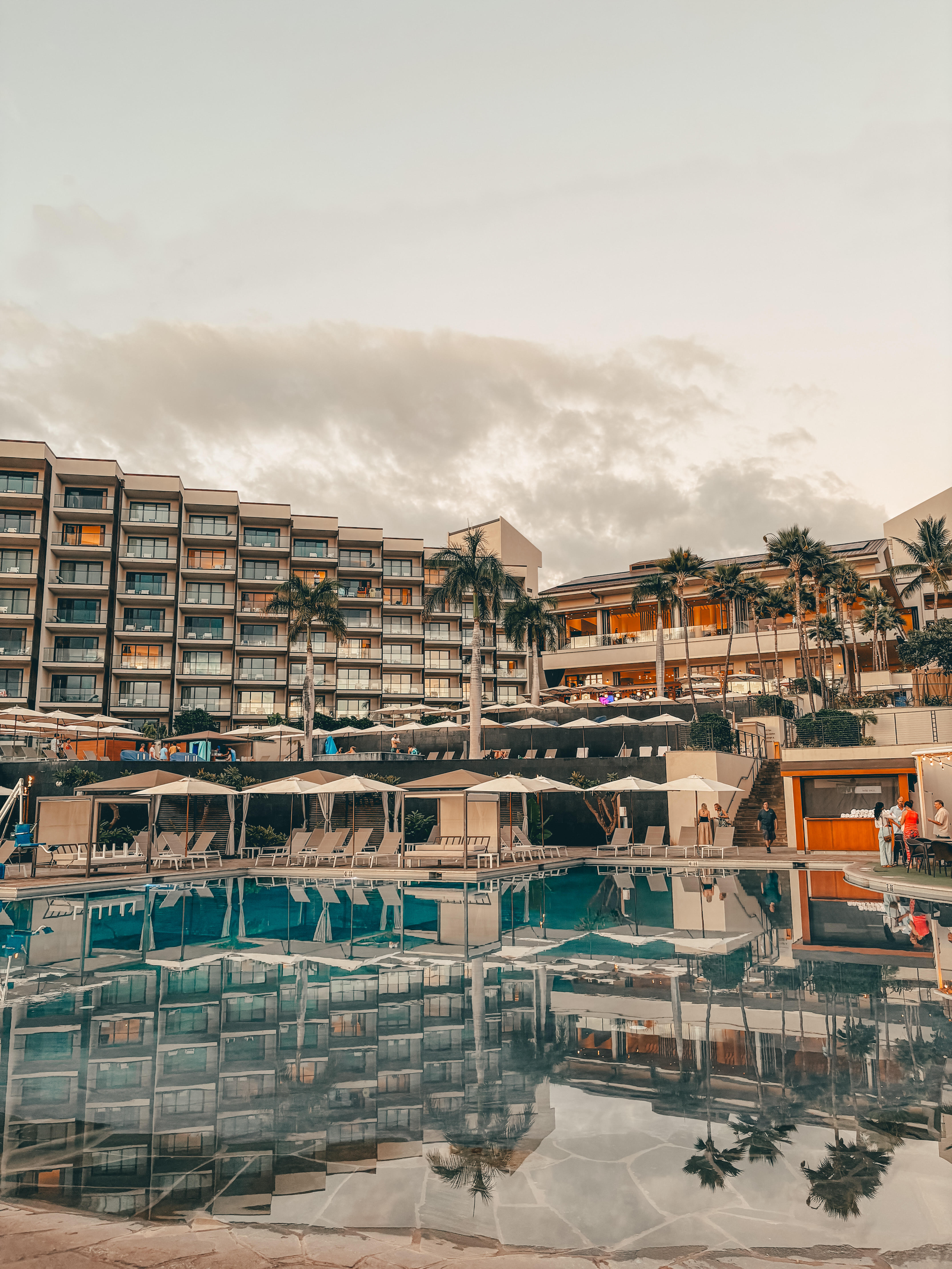 A luxurious resort with modern architecture, featuring a large infinity pool reflecting the surrounding multi-story buildings, palm trees, and sky at dusk. Poolside lounge chairs and cabanas line the water's edge, while warm lights from the hotel balconies and an outdoor bar add a cozy ambiance. A few guests can be seen enjoying the resort, creating a relaxed and upscale tropical atmosphere.