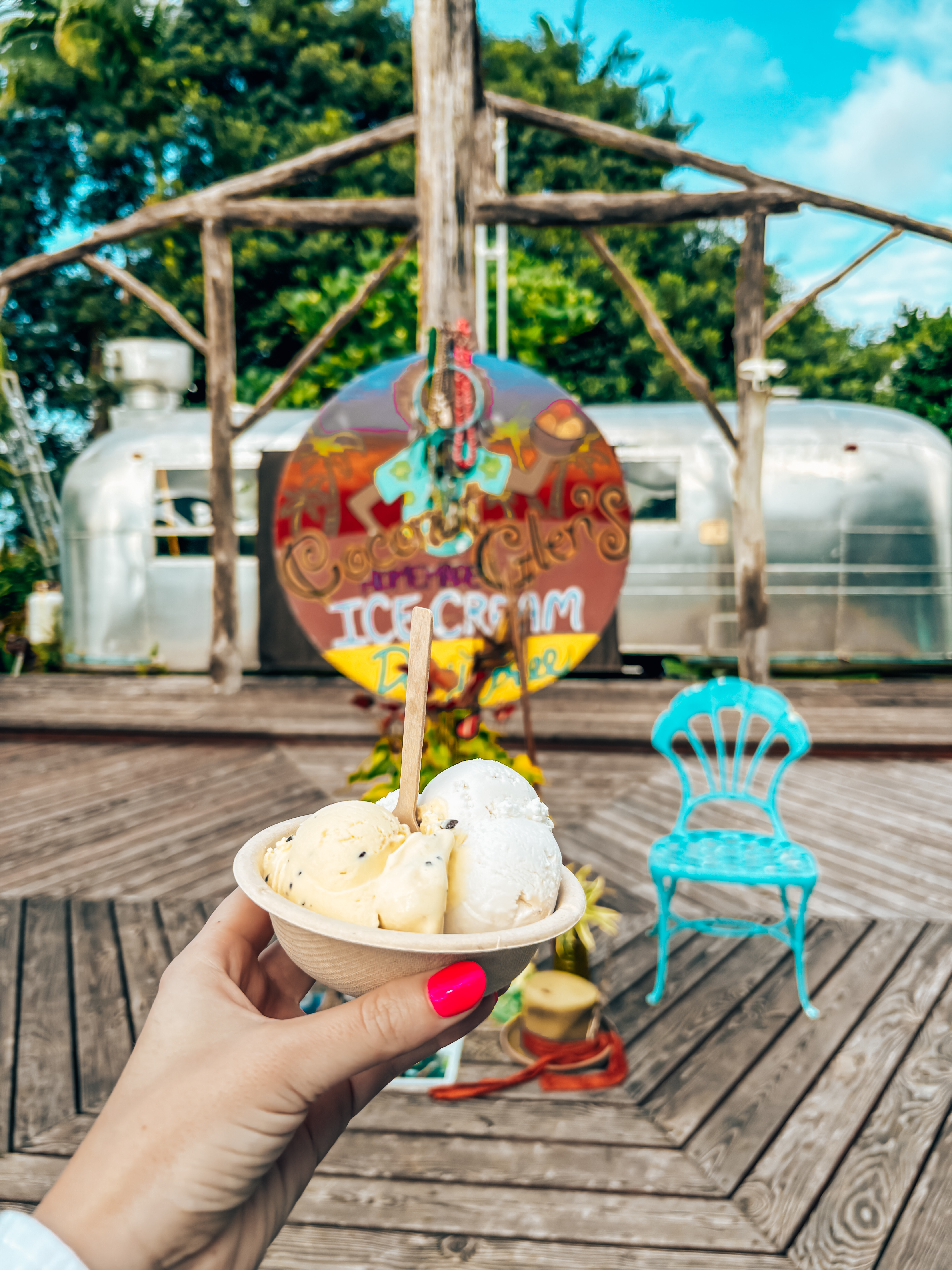 A hand with bright pink nail polish holds a small biodegradable bowl filled with three scoops of coconut-based ice cream, with a wooden spoon sticking out. In the background, a colorful sign reading "Coconut Glen's Ice Cream" is displayed in front of a vintage silver Airstream trailer, with rustic wooden beams and a bright blue chair on a wooden deck. The tropical setting, lush greenery, and vibrant decor add to the charming roadside atmosphere.