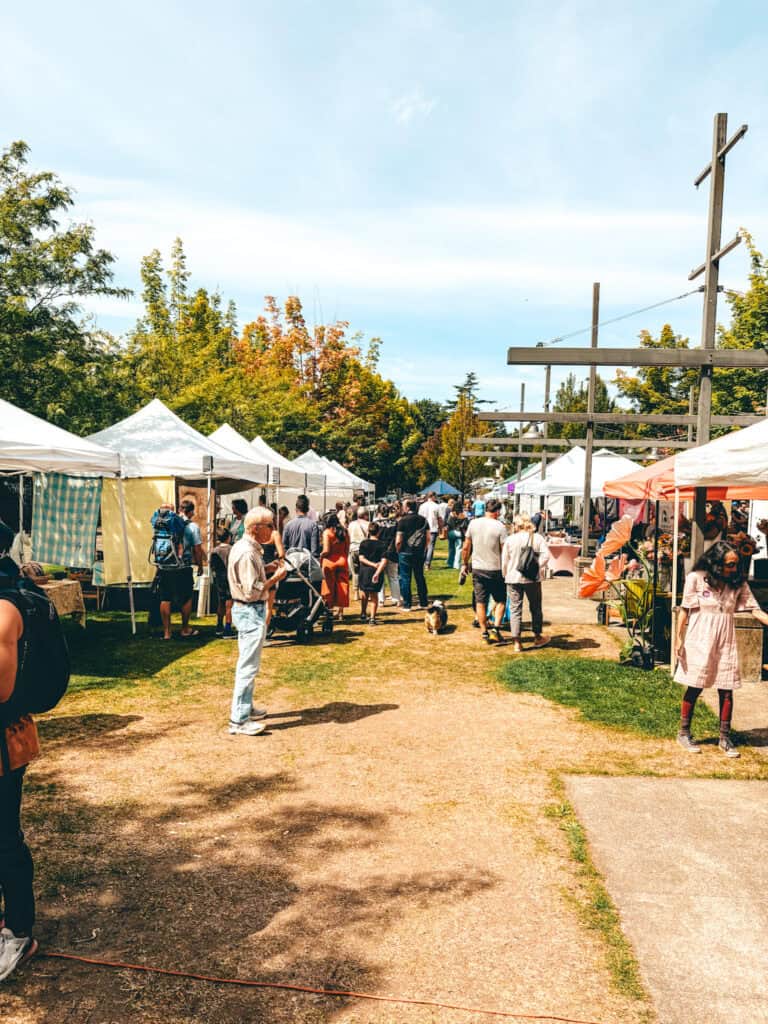 Outdoor farmers market on a sunny day with rows of white and colorful vendor tents lining a grassy path. A crowd of people, including families and shoppers with dogs, browse local goods and produce under the shade of green and autumn-tinged trees.