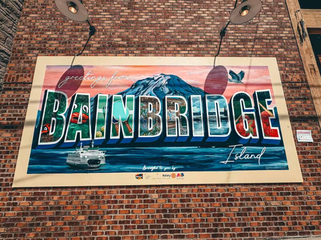 Mural on a brick wall reading "Greetings from Bainbridge Island" in large block letters filled with Pacific Northwest imagery like salmon, mountains, a ferry, and a bald eagle. The scene includes a backdrop of Mount Rainier at sunset and a Washington State ferry on the water below the text.