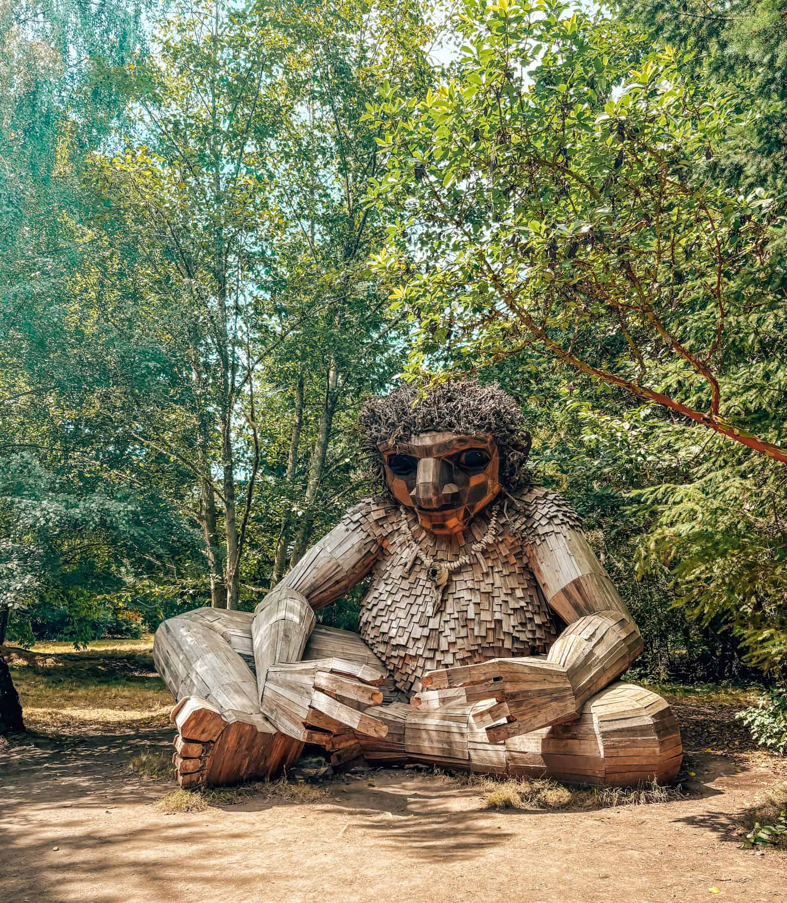 Large wooden troll sculpture seated cross-legged in a forest clearing, made from reclaimed wood with intricate details including textured hair, facial features, and layered wood scales on the torso. Surrounded by lush green trees, the troll blends into its natural setting while exuding a whimsical, mythical presence.