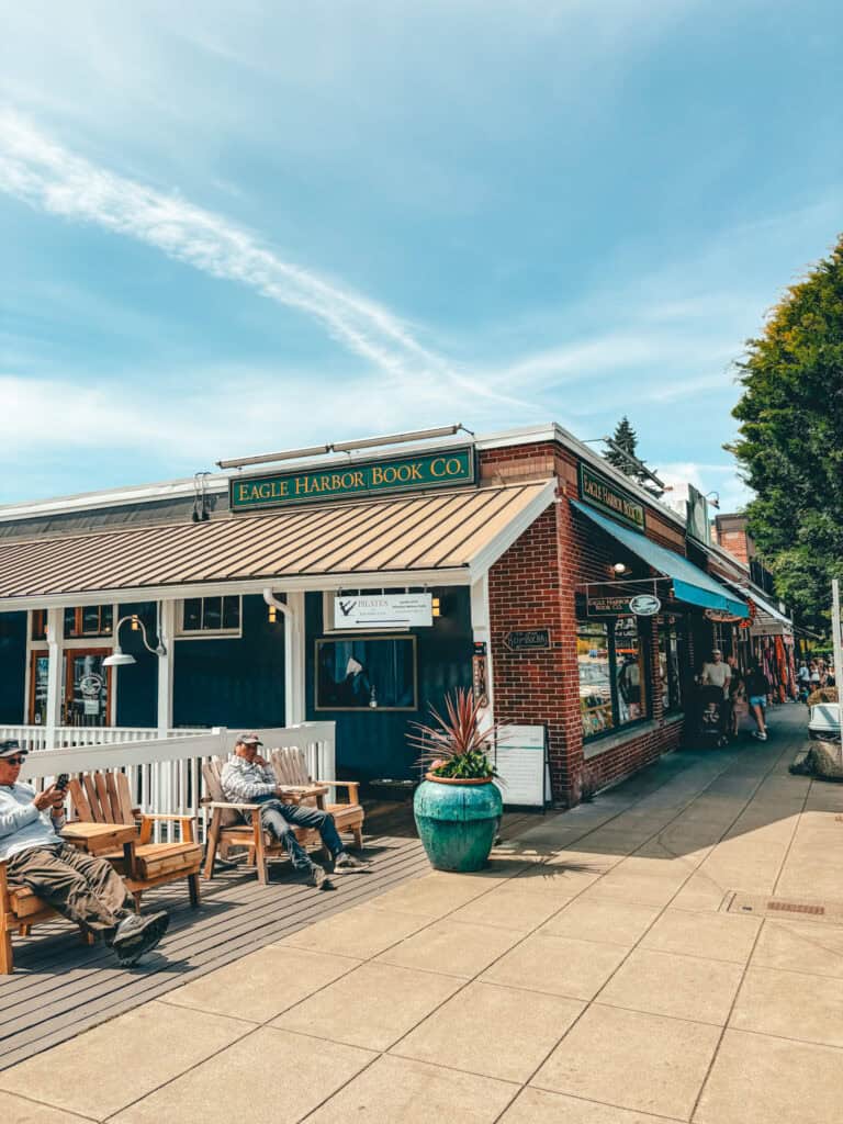 Street view of Eagle Harbor Book Co., a charming independent bookstore on Bainbridge Island with a green and gold sign above the entrance. Two men relax on wooden benches outside under a sunny blue sky, while pedestrians browse shops along the sidewalk.
